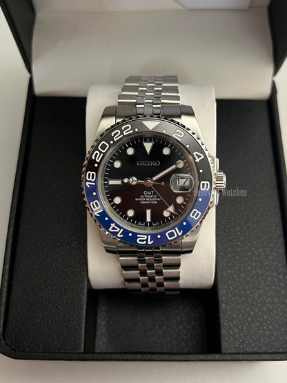 GMT (Batman) Seiko NH34 GMT automatic mod watch. A front view of the watch in a box featuring a black dial and blue and black bezel on a silver bracelet.